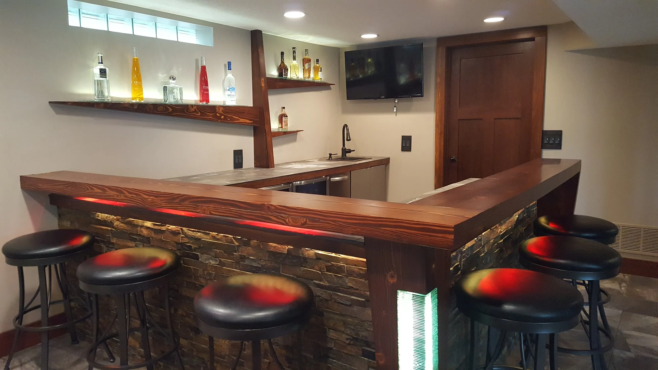Basement bar with a stacked stone front, wood bar top, and LED-lit rail